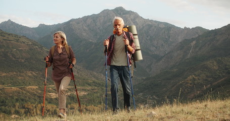 Positive mature caucasian couple trekking in mountains with backpacks, enjoying their adventure 4k