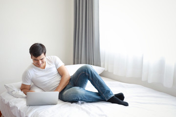 portrait of handsome caucasian in white shirt and blue jean using computer on bed in bedroom with relaxation and smiling face