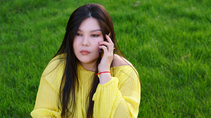 Focused plus size pan-asian girl sitting on the grass and talking by the smartphone in a summer...