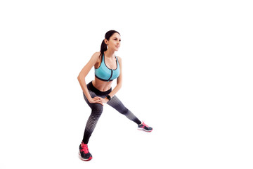  A young woman in comfortable sportswear (leggings and top) is smiling charmingly and doing wide lunges to the sides with her legs on an isolated white background.