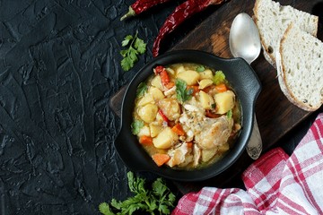 Chicken stew with potatoes and vegetables. in a black pan for serving dishes on a dark background surrounded by spices. meat and vegetables are pre-fried and then stewed. top view. copy space. 