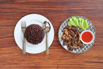 Deep fried beef served with sliced cucumber and sweet chili sauce served with cooked Thai black jasmine rice berry on wooden board background.