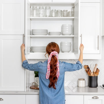 A Woman Looking In A Farmhouse Kitchen Cabinet Full Of White Dishes