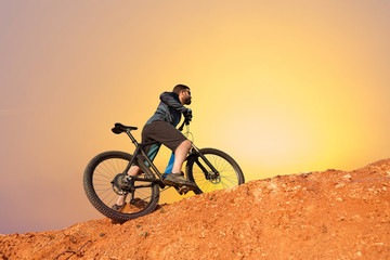 Cyclist in shorts and jersey on a modern carbon hardtail bike with an air suspension fork rides off-road on the orange-red hills at sunset evening in summer