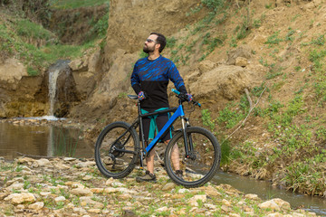 Obraz premium Cyclist in shorts and jersey on a modern carbon hardtail bike with an air suspension fork stands on the rocky shore of a mountain river