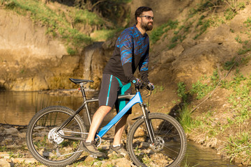 Obraz premium Rowerzysta w krótkich spodenkach i koszulce na nowoczesnym rowerze typu hardtail z włókna węglowego z widelcem pneumatycznym stoi na skalistym brzegu górskiej rzeki