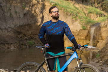 Cyclist in shorts and jersey on a modern carbon hardtail bike with an air suspension fork stands on the rocky shore of a mountain river