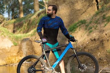 Fototapeta premium Cyclist in shorts and jersey on a modern carbon hardtail bike with an air suspension fork stands on the rocky shore of a mountain river