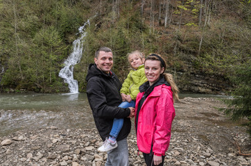Naklejka na ściany i meble Young happy family: mom, dad and daughter on the background of a mountain waterfall