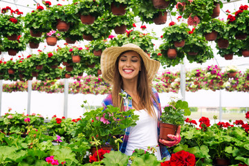 Beautiful attractive woman florist with hat walking through greenhouse holding potted flowers and checking plants. Happy lifestyle.