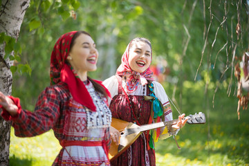 Two young attractive women in traditional russian clothes singing in the forest. One of them playing balalaika - obrazy, fototapety, plakaty