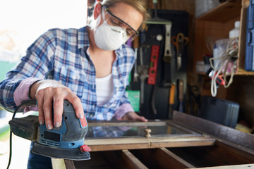 Mature Woman Upcycling Furniture In Workshop At Home Using Electric Sander