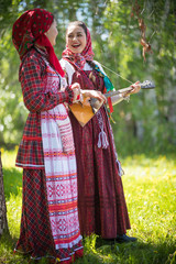 Two young woman in traditional russian clothes standing in the forest and look at each other. One...