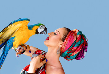 Portrait of young attractive woman in african style holding ara parrot on her hand onblue background