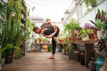 A man practices yoga asanas on a veranda in a greenhouse with flowers.