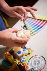 Close-up of seamstress taking pin from pillow to use it in sewing while standing near the table with pattern and colorful threads