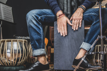A stylish musician in denim and double monk shoes plays the Cajon, a Peruvian drum used commonly...