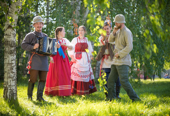 People in traditional Russian clothes are standing in the woods and talking - one of them plays the...