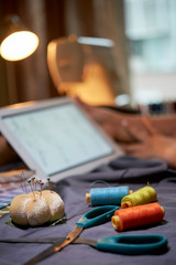 Close-up of scissors, pins and colorful threads with fabric on the table in workshop and with digital tablet in the background