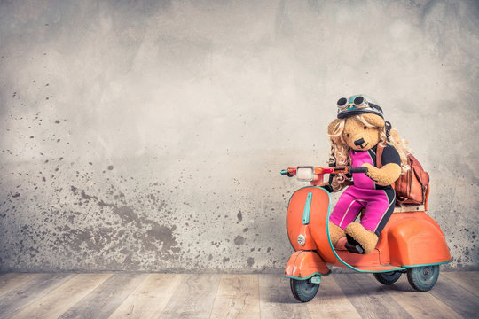 Retro Teddy Bear With Long Hair Toy In Helmet With Goggles And Backpack On Children's Pedal Scooter From 60s Front Concrete Wall Background. School Girl Kid Concept. Vintage Old Style Filtered Photo