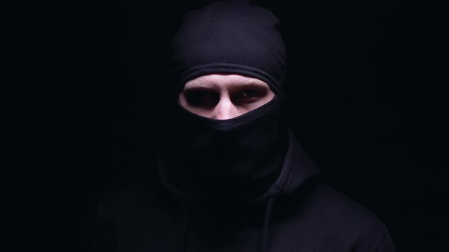 Criminal in dark balaclava angrily looking into camera against black background