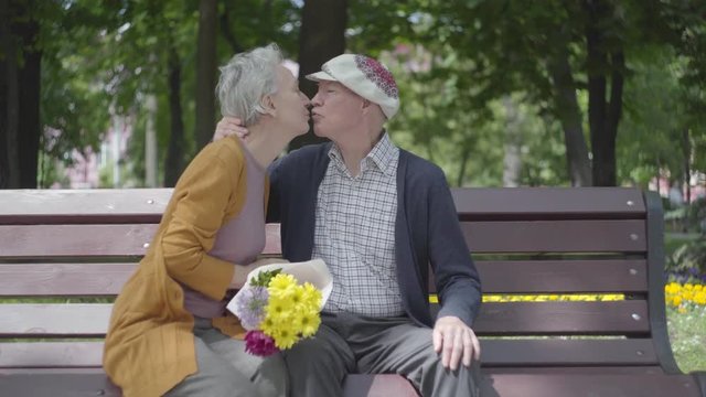 Old man kissing his woman with flowers on the banch in a spring park