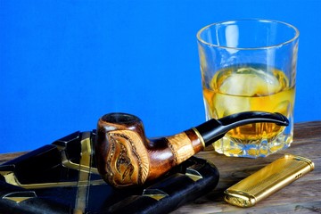 Smoking pipe with an ornament in an ashtray, a lighter and alcohol with ice. A Smoking pipe for the ritual Smoking of tobacco. Strong alcoholic drink with ice on a wooden table top - whiskey or Bourbo