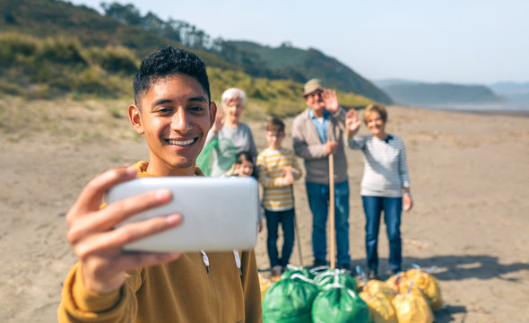 Young man taking selfie with mobile to group of volunteers after cleaning the beach. Selective focus on guy in foreground