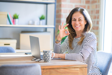 Middle age senior woman sitting at the table at home working using computer laptop doing ok sign with fingers, excellent symbol