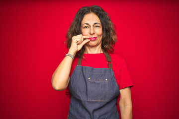 Middle age senior woman wearing apron uniform over red isolated background mouth and lips shut as...