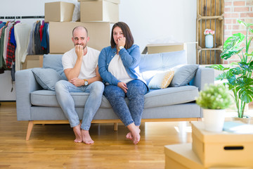 Fototapeta na wymiar Young couple sitting on the sofa arround cardboard boxes moving to a new house looking stressed and nervous with hands on mouth biting nails. Anxiety problem.