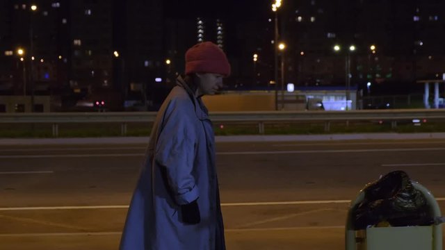 homeless person walks along highway against background of night city. bum digging in trash urn. Tramp drinks drink from paper cup.