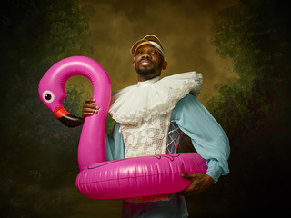 Young man as a medieval grandee or nobleman in a swimring as a pink flamingo on dark studio background. Portrait in retro costume. Human emotions, comparison of eras and facial expressions concept.