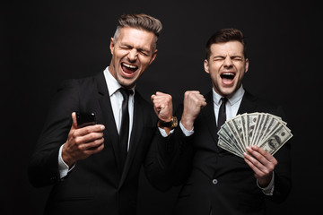 Excited happy two business men posing isolated over black wall background using mobile phone...