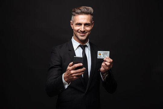 Business man posing isolated over black wall background using mobile phone holding credit card.