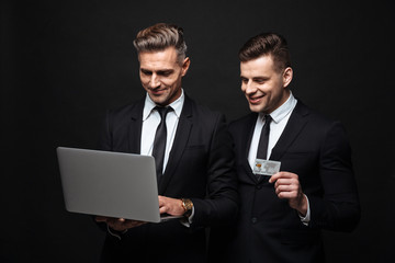 Handsome excited happy two business men posing isolated over black wall background using laptop computer holding credit card.