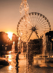 Unrecognizable Kid Playing in Water Fountain and Ferris Wheel in Sunset in France.