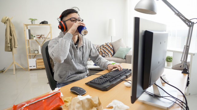 Asian Weird Guy Stay At Home Indoorsy In Summer Break All Day In Dirty Apartment. Young Man Drinking Can Of Beverage While Playing Online Game On Computer On Messy Desk. Lazy Male Lifestyle Concept.