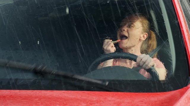 woman drives a car and does makeup at the same time. girl behind the wheel of a car paints her lips creating an emergency on the road. It is raining outside.