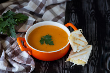 Pumpkin carrot cream soup in an orange plate with cheese pita on a checkered tablecloth with wooden background. Homely very tasty dinner and lunch. Family meal. Delicious main course. Cozy food served