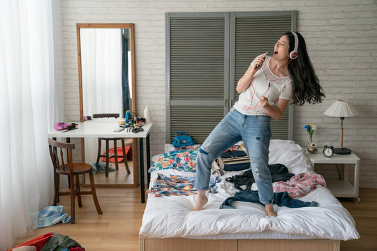 Young asian woman listening music from smart phone via headphones and singing along on bed dancing having fun get crazy. female prepared clothes in suitcase relax in break time at home bedroom.