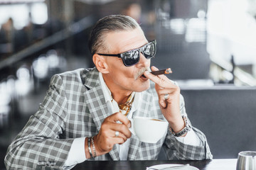 Businessman with suspicious face drinks caffe  beverage and smokes cigarette on summer terrace