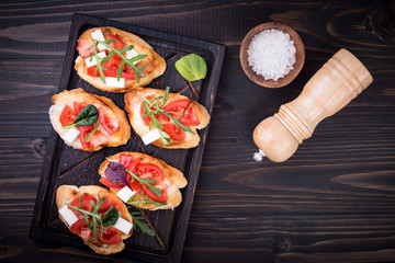 Bruschetta with tomatoes, mozzarella cheese and herbs on a cutting board in a dark background, top view