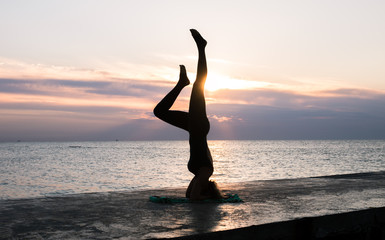 unrecognizable woman with beautiful body doing yoga headstand at sunrise on the sea, silhouette of yoga poses