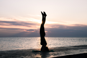unrecognizable woman with beautiful body doing yoga headstand at sunrise on the sea, silhouette of yoga poses