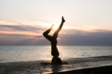 unrecognizable woman with beautiful body doing yoga headstand at sunrise on the sea, silhouette of yoga poses