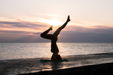 unrecognizable woman with beautiful body doing yoga headstand at sunrise on the sea, silhouette of yoga poses