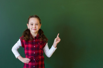 Happy schoolgirl preschool girl in plaid dress standing in class near a green blackboard. Concept of school education