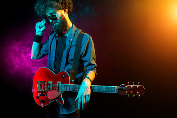 Obraz premium Young hipster man with curly hair with red guitar black sunglasses in neon lights. Rock musician is playing electrical guitar.
