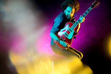 Young hipster man with curly hair jump with red guitar in neon lights. Rock musician is playing...